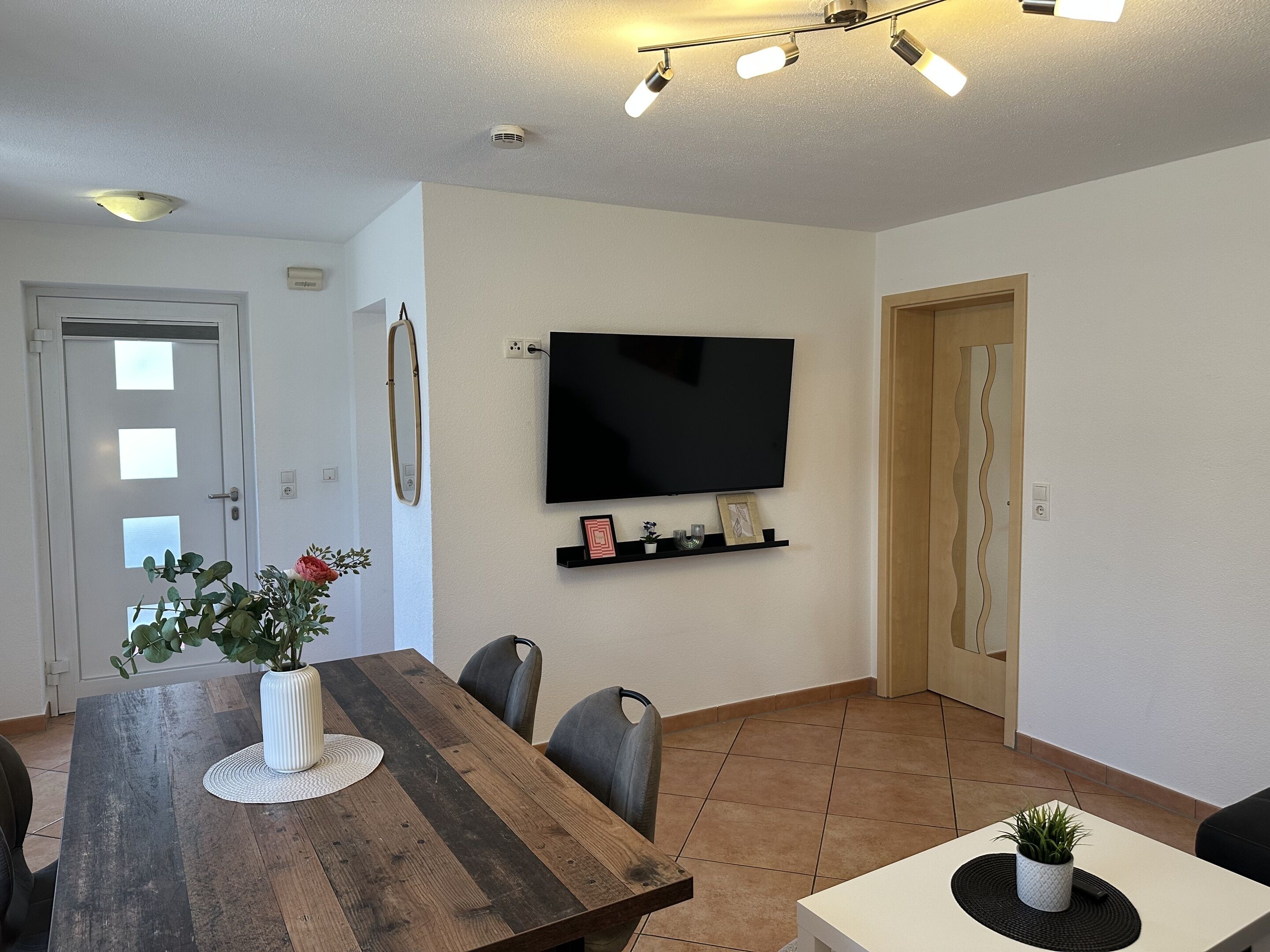 Un comedor con una mesa de madera, sillas, un televisor montado en la pared y una puerta.