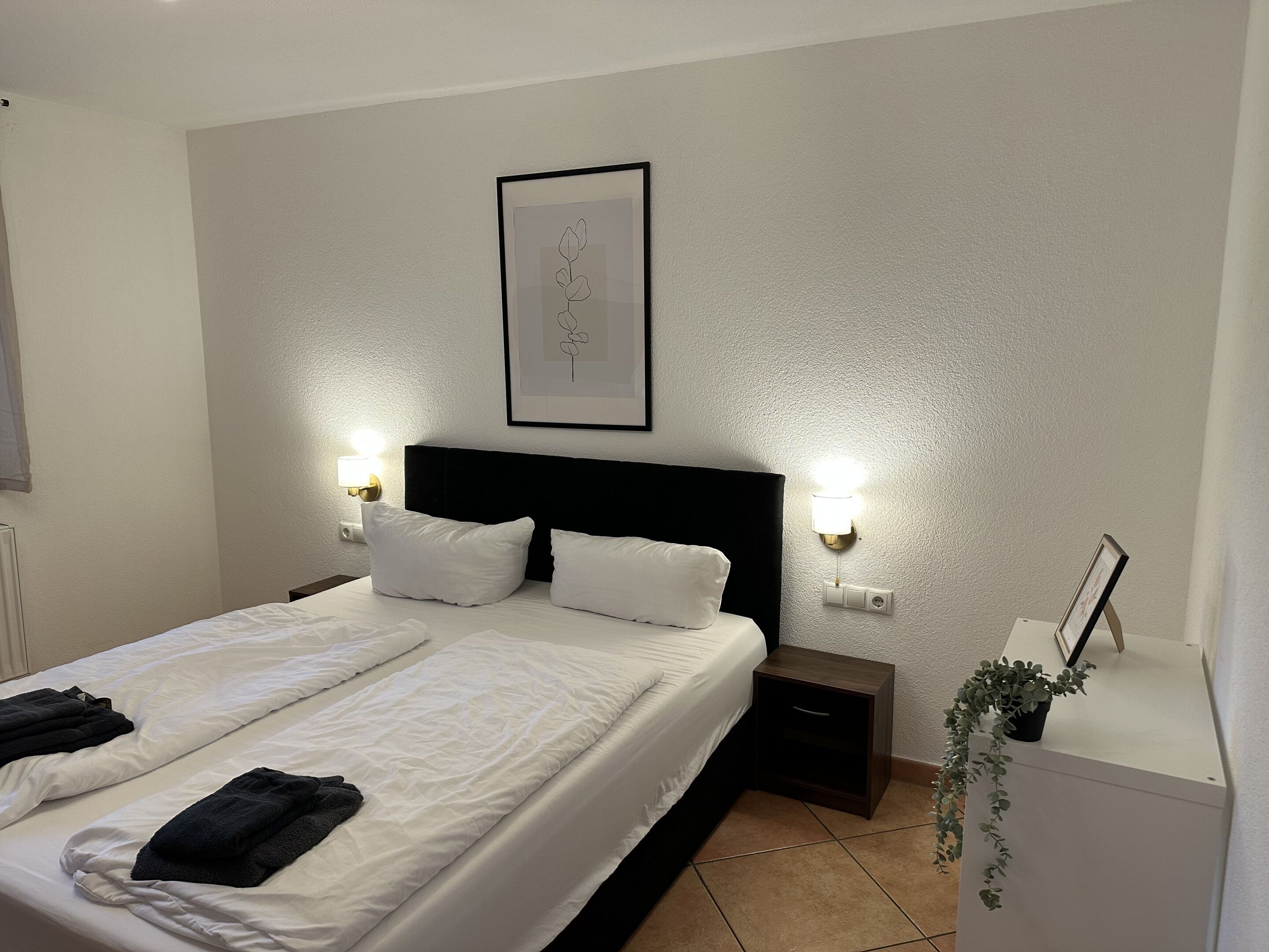 A hotel room with a large bed, two bedside tables, a nightstand, a framed picture on the wall, and a potted plant.