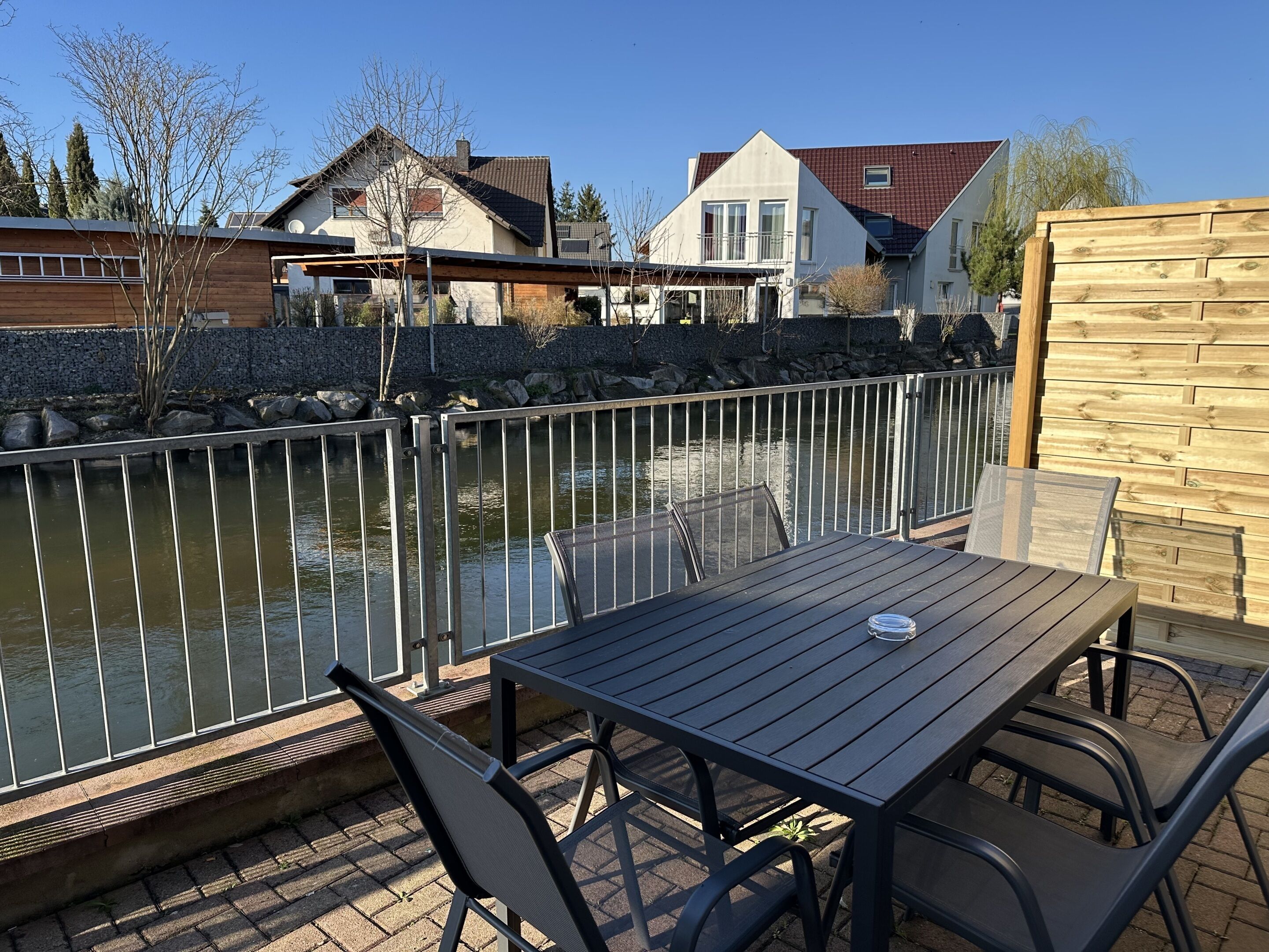 Eine Terrasse mit Tisch und Stühlen, die auf ein Gewässer mit Häusern im Hintergrund blickt.