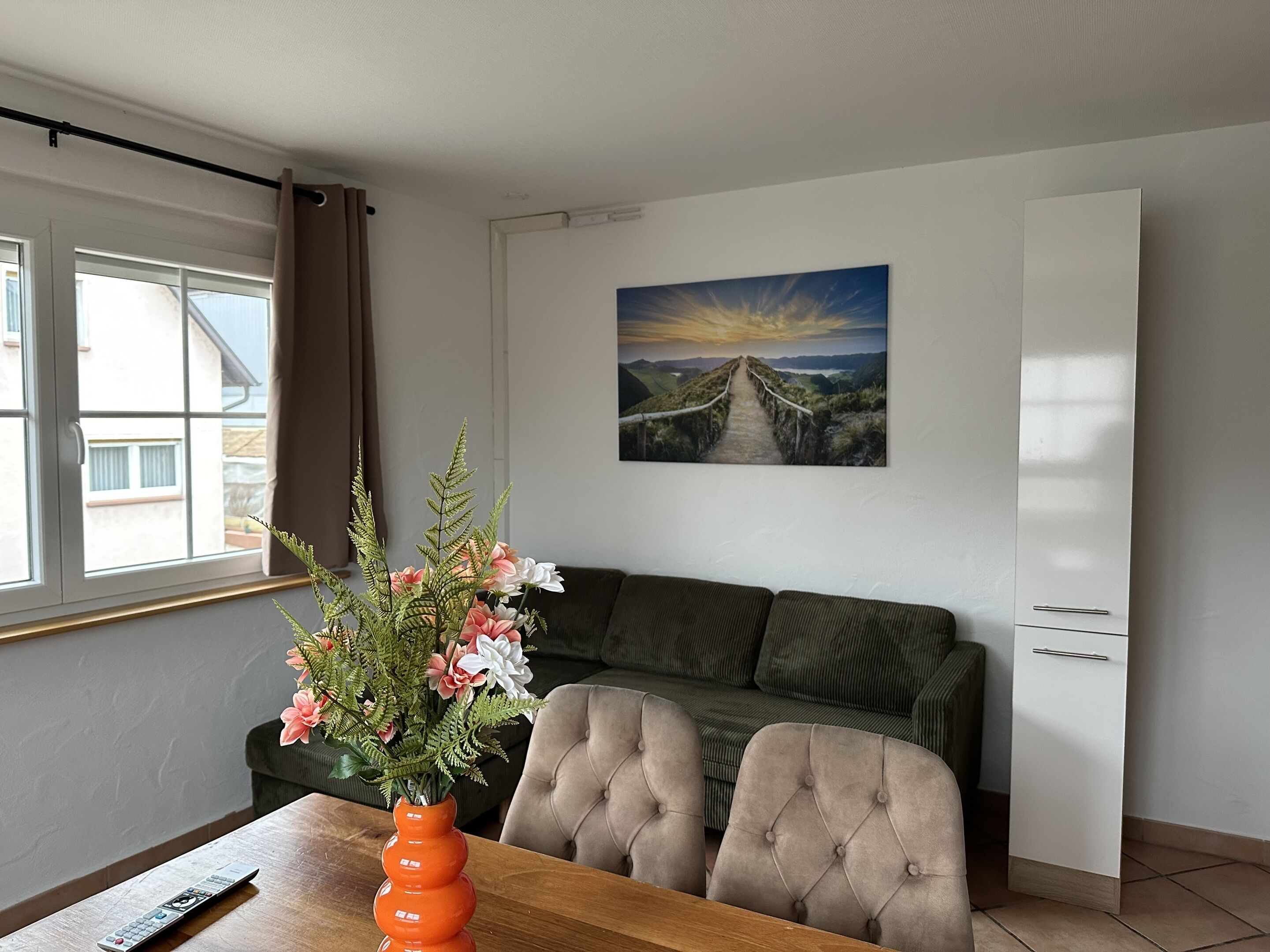 Un área de comedor con una mesa de madera, un florero con flores, un control remoto, un sofá y un cuadro de un atardecer en la pared.