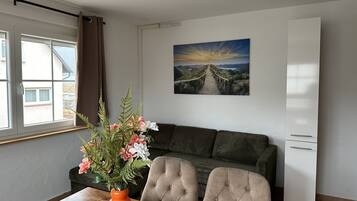 Un área de comedor con una mesa de madera, un florero con flores, un control remoto, un sofá y un cuadro de un atardecer en la pared.