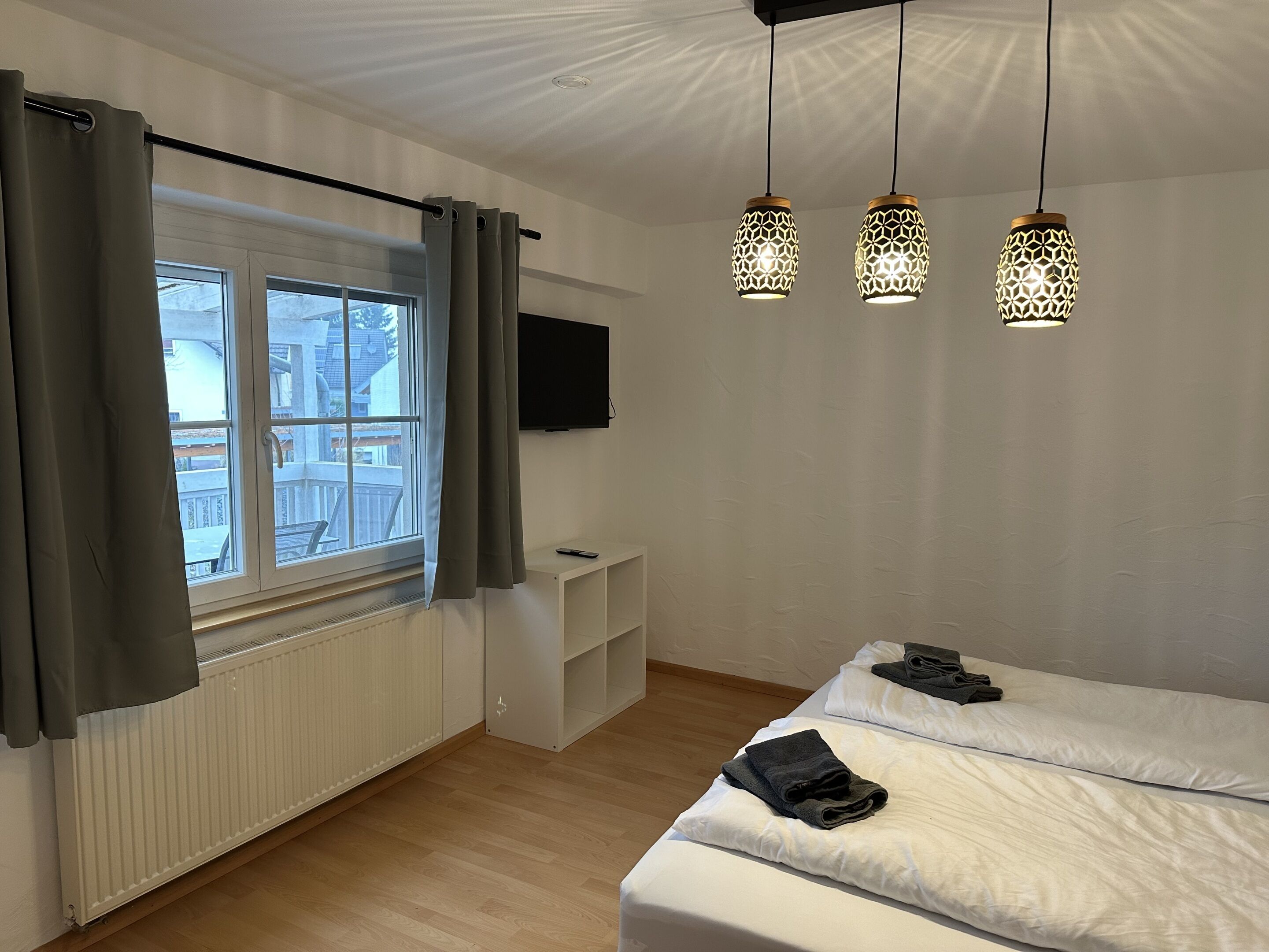 Ein Hotelzimmer mit Bett, einem Fenster mit Vorhängen, einem an der Wand montierten Fernseher und drei Pendelleuchten.