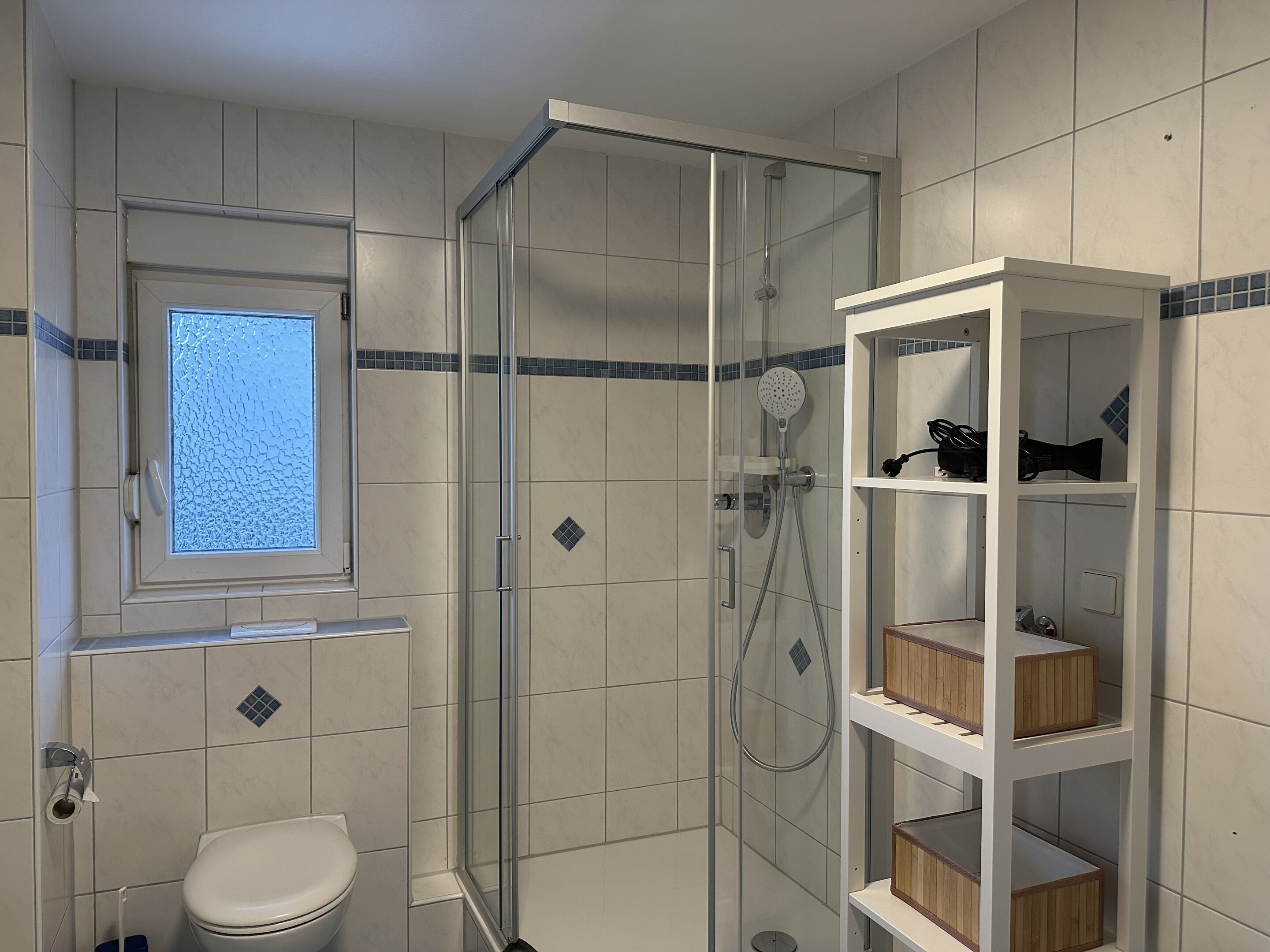 Een badkamer met een douche in een glazen cabine, een toilet, een raam en een houten plankunit.