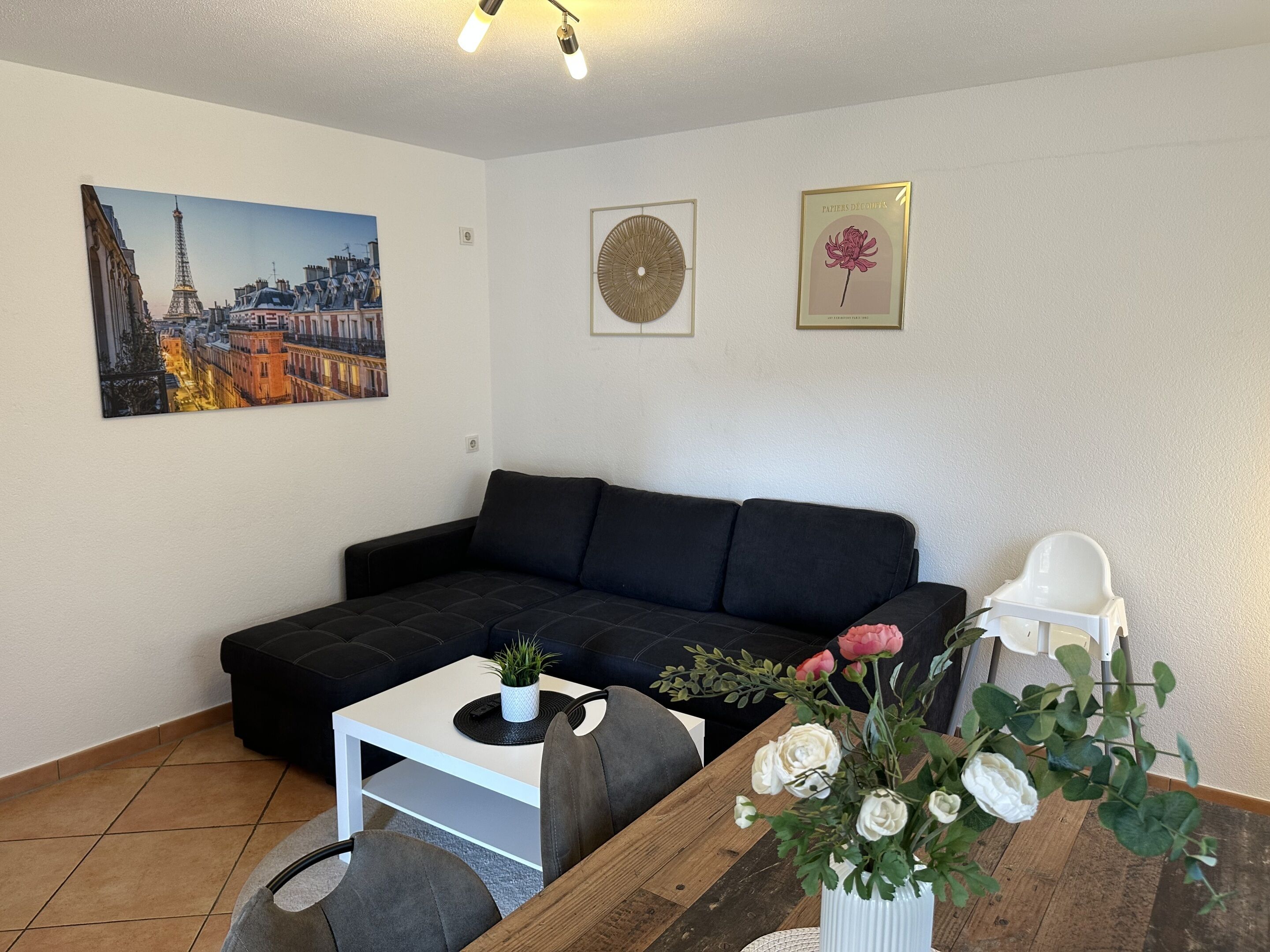 Una sala de estar con un sofá negro, una mesa de centro blanca, una pequeña planta en maceta, una mesita de madera con un jarrón de flores, y dos cuadros en la pared.
