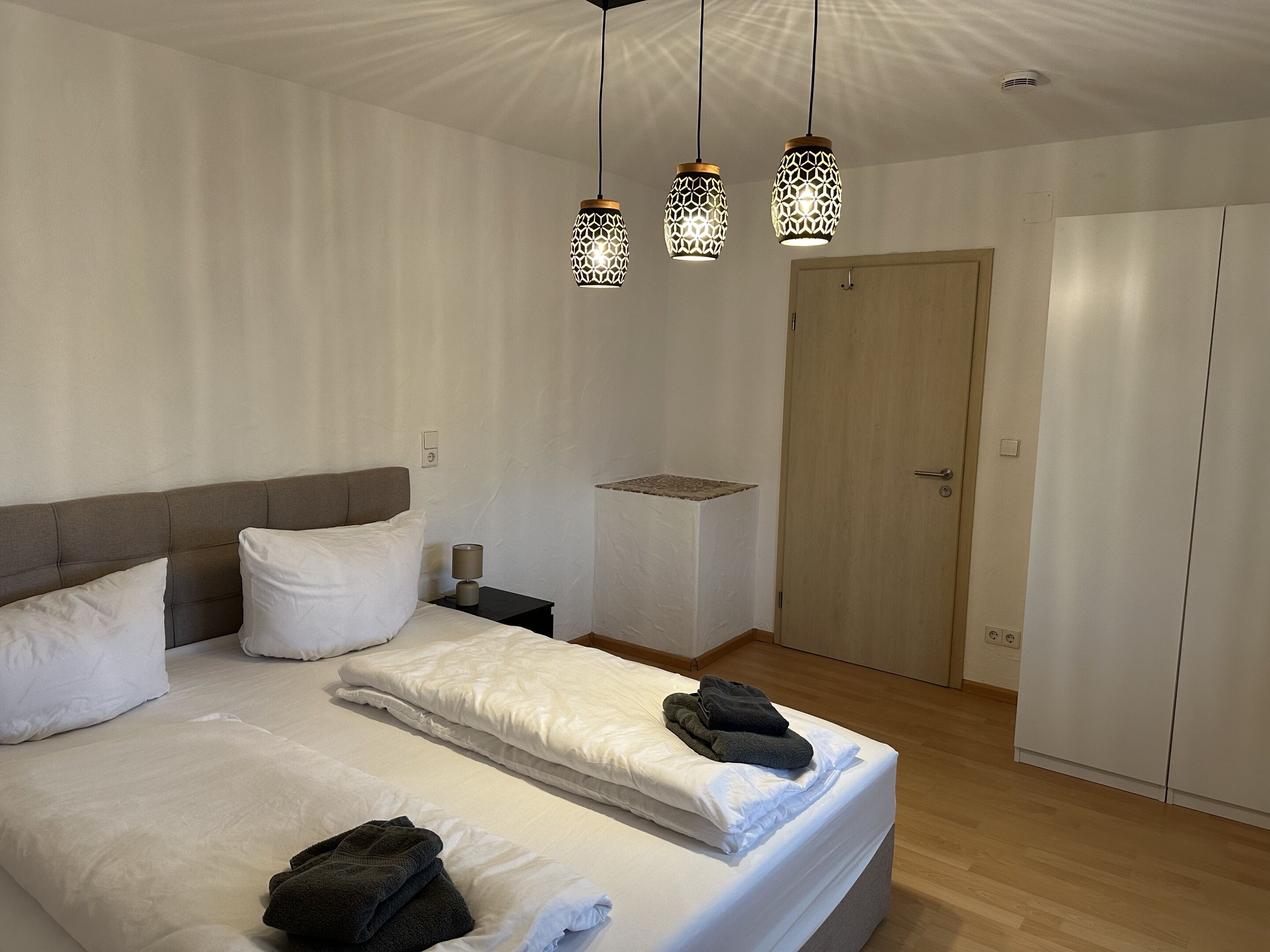 A neatly made bed with white linens, a bedside table with a lamp, and a wardrobe.