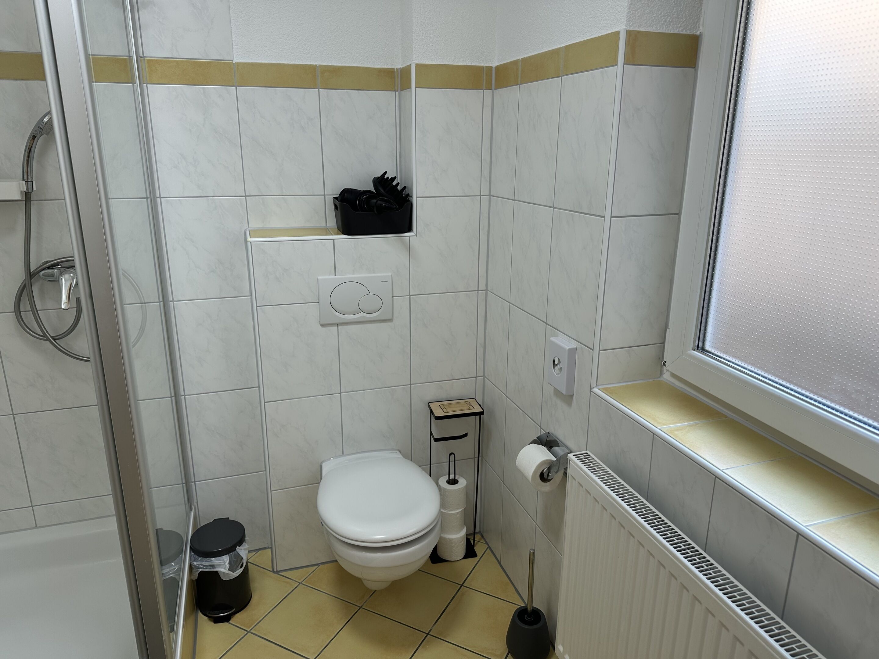 Een kleine badkamer met toilet, prullenbak, toiletborstel, radiator en raam.