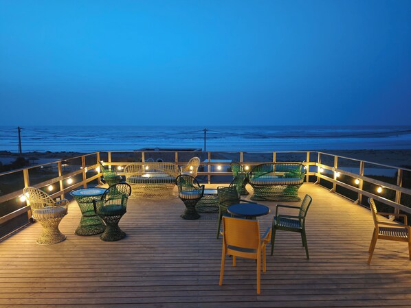 A wooden deck with wicker furniture and string lights, overlooking a beach at dusk. - The Mogador's Oasis (Akermoud)