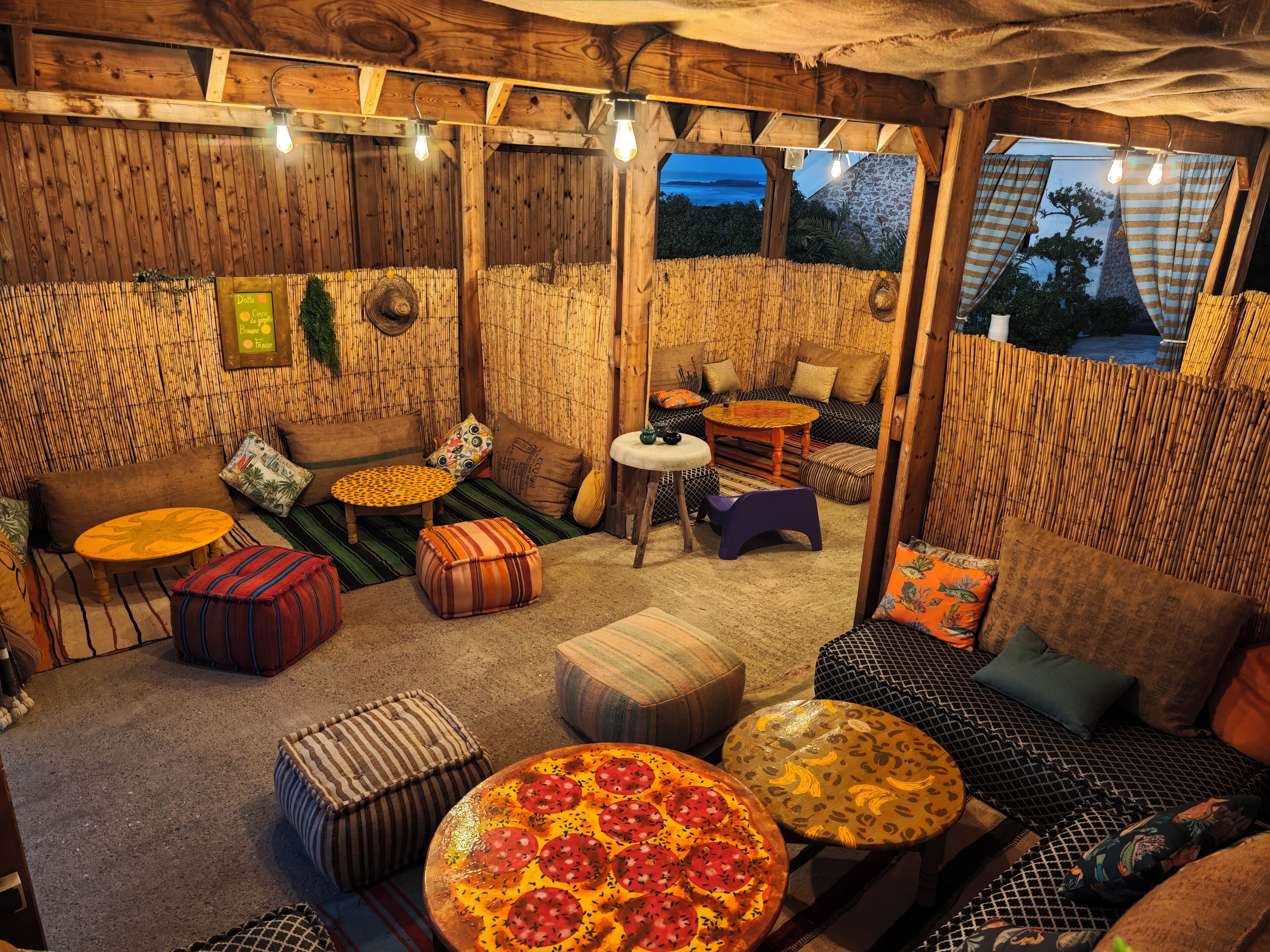 A cozy, rustic interior with wooden walls, a large pizza on a round table, and a view of the sea through a window.