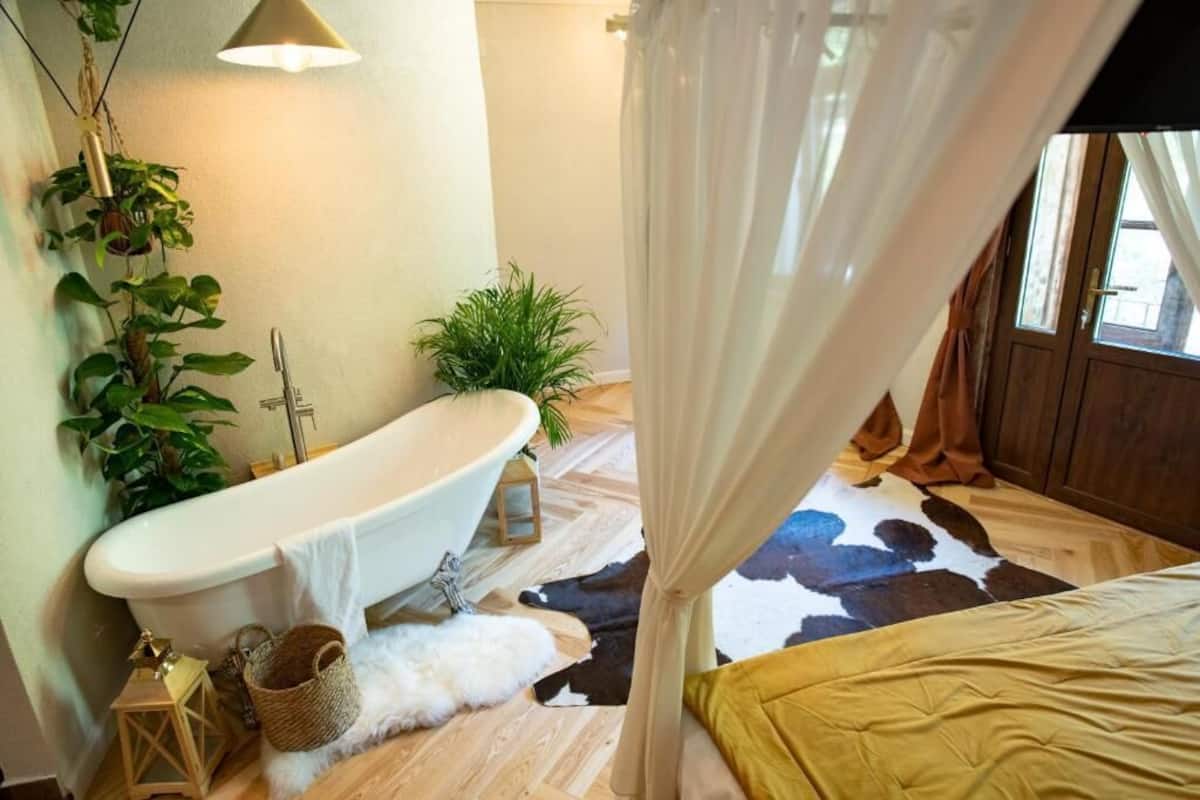 Un dormitorio con un ventanal grande, una cama con colcha amarilla, un felpudo de piel blanca, un tapiz de cuero de vaca, una bañera y una planta colgante.