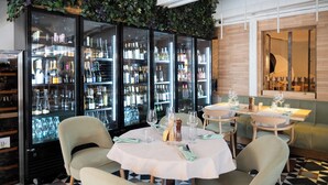 Interior de restaurante con una zona de bar que incluye refrigeradores con puertas de vidrio llenos de botellas, un área de comedor con mesas para dos y un banco acolchado verde.