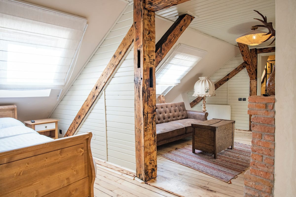 Un dormitorio tipo loft con techo inclinado, vigas de madera, una cama, un sofá, una mesita y una lámpara.