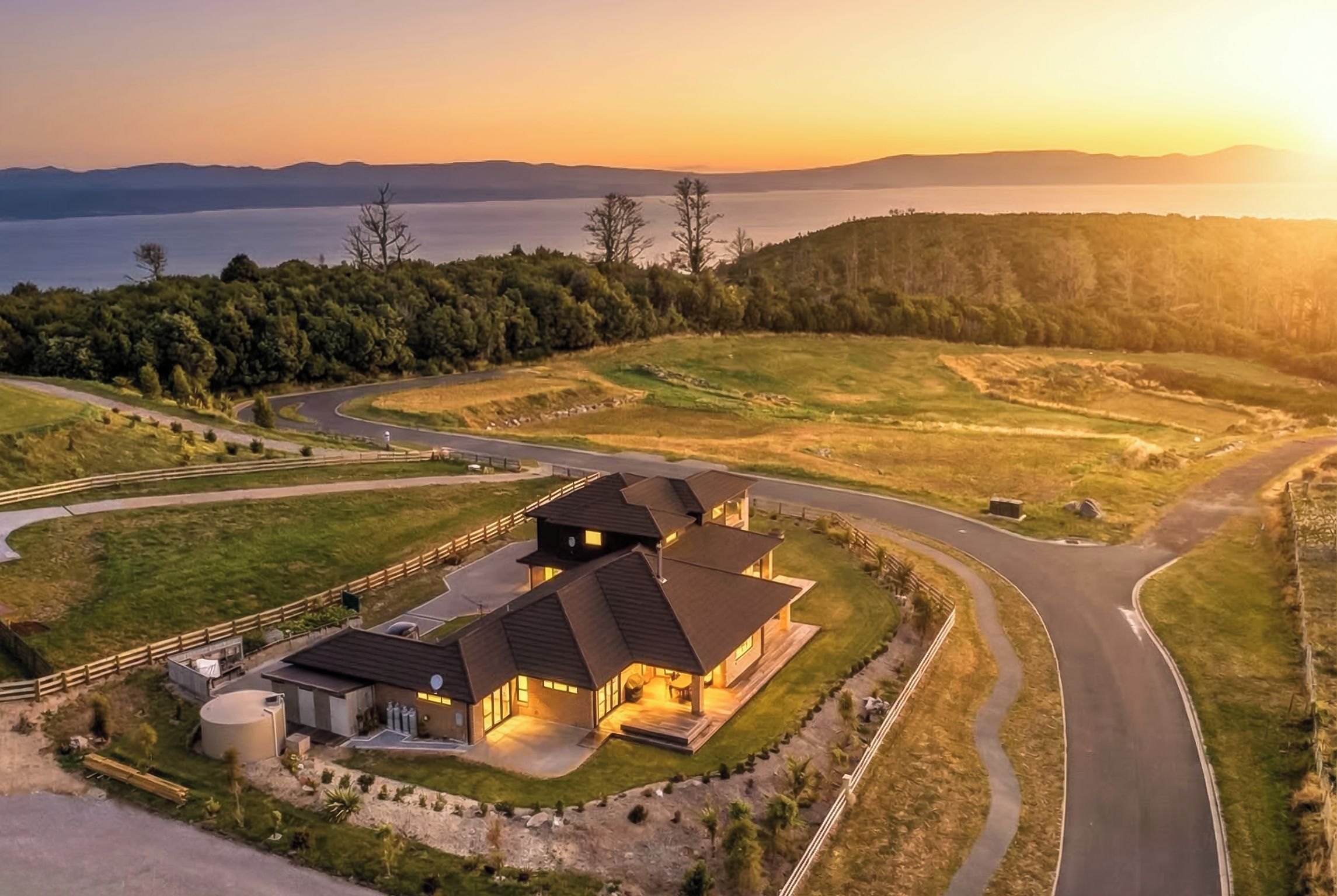 A modern house with a driveway, surrounded by a landscaped garden, with a view of a lake and hills in the background.