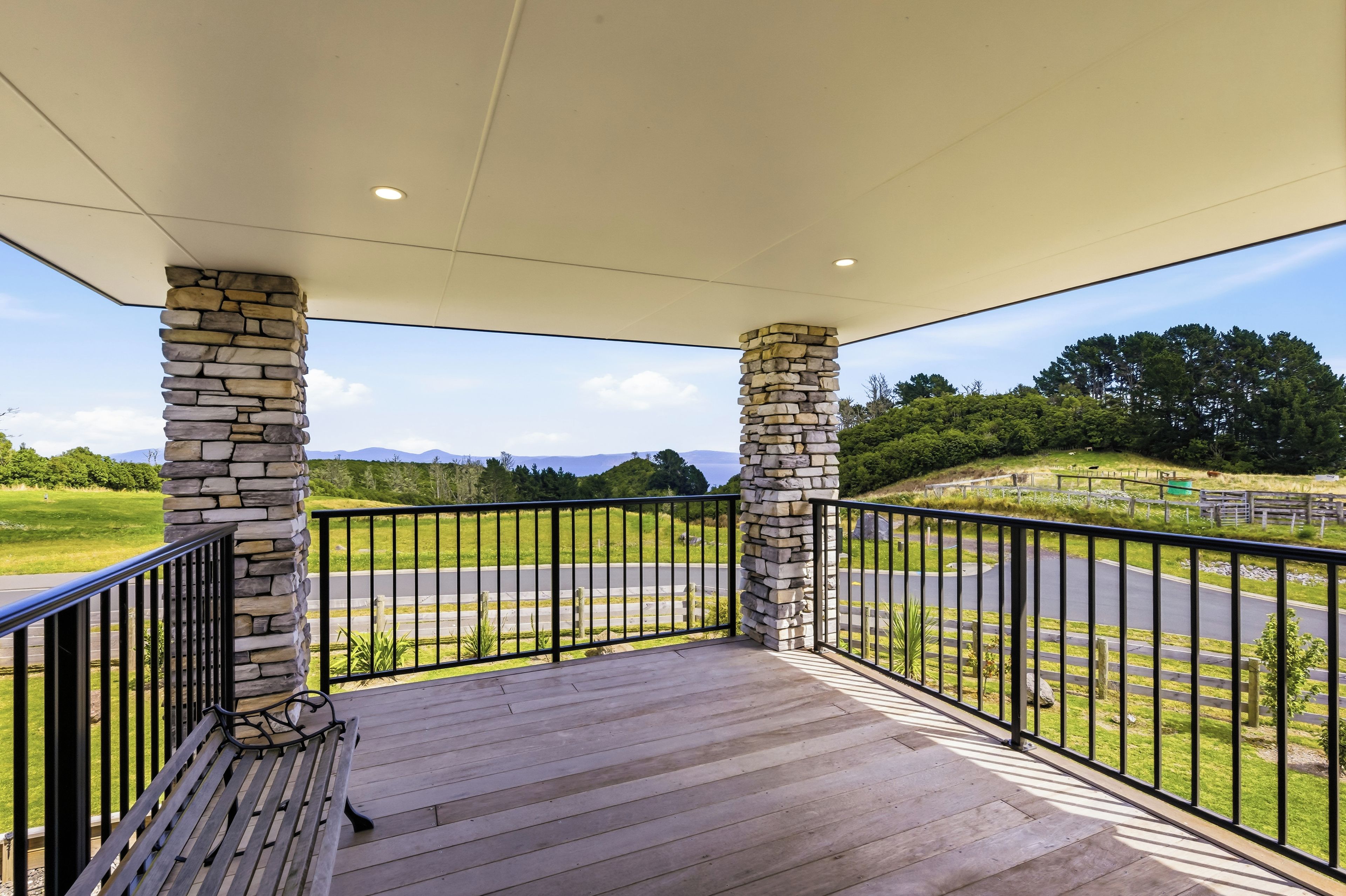 A covered outdoor area with stone pillars, a wooden deck, and a view of a road and greenery.