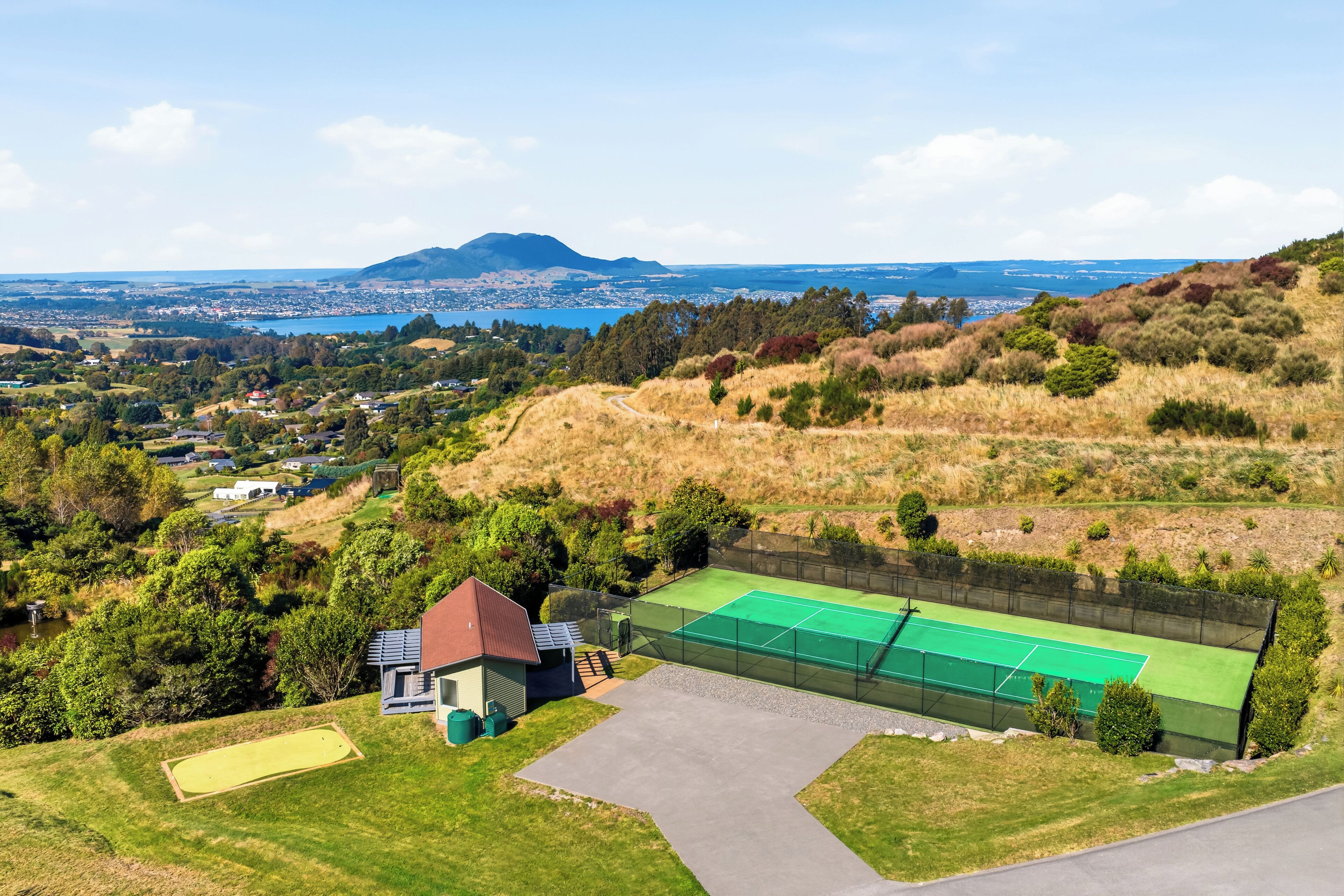 A tennis court with a net, a building with a red roof, and a view of a town and mountains in the distance.
