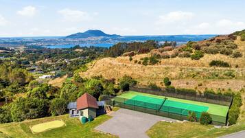 A tennis court with a net, a building with a red roof, and a view of a town and mountains in the distance.