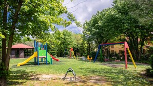 Un terrain de jeux avec des toboggans et des balançoires, entouré d’arbres, et un bâtiment visible au loin.