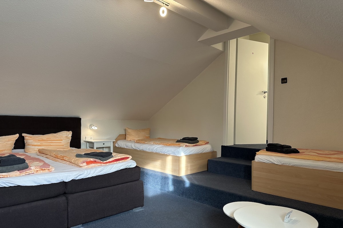 Una camera d'albergo moderna con un letto grande, un letto più piccolo, una poltrona letto, una scrivania e un tavolino.
