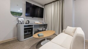 Una habitación de hotel moderna con sofá, una pequeña mesa redonda, un escritorio con televisor de pantalla plana, una cafetera y un hervidor.