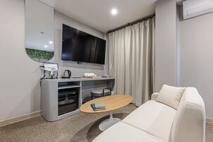 Una habitación de hotel moderna con sofá, una pequeña mesa redonda, un escritorio con televisor de pantalla plana, una cafetera y un hervidor.