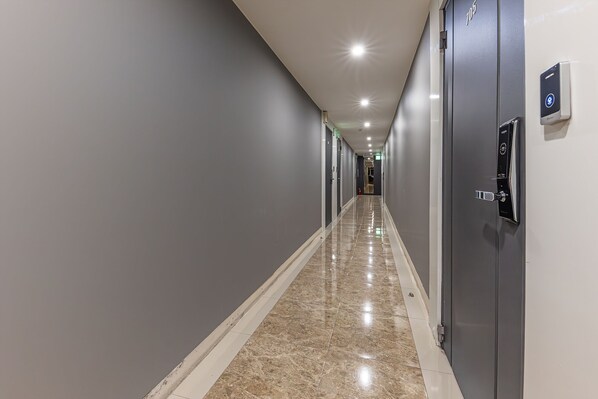 A long, narrow hotel corridor with grey walls, a marble floor, and a series of doors on the right side. - hotelyello Guri (Guri)