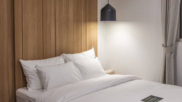 Una cama bien hecha con sábanas y almohadones blancos, un panel de pared de madera, una lámpara colgante negra y una mesita de noche con un objeto pequeño encima.