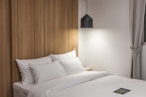 Una cama bien hecha con sábanas y almohadones blancos, un panel de pared de madera, una lámpara colgante negra y una mesita de noche con un objeto pequeño encima.