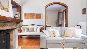A living room with a fireplace, a grey sofa with multiple cushions, framed pictures on the wall, and a wooden archway leading to another room.