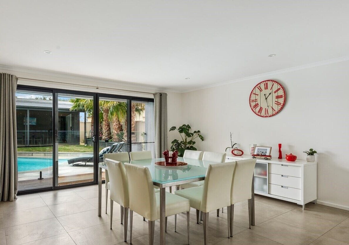 Un espace repas moderne avec une table en verre et des chaises blanches, un meuble blanc avec des tiroirs et une grande horloge murale.