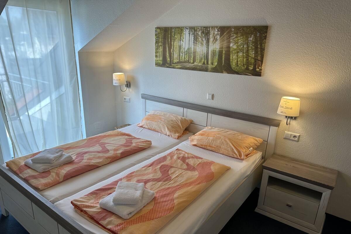 A double bed with two pillows, a bedside table, a wall-mounted lamp, and a forest-themed painting.