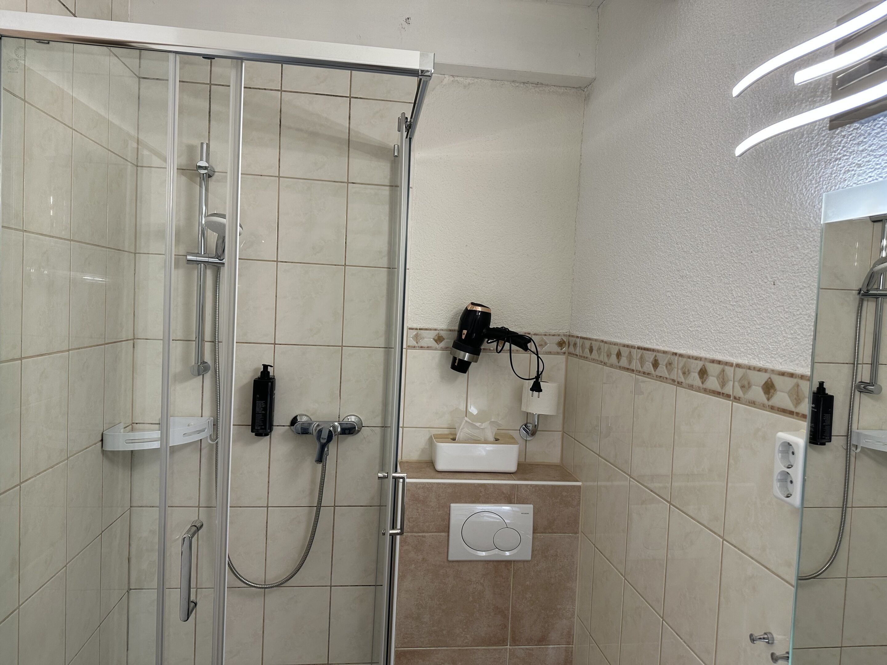 Une douche avec une porte en verre, un pommeau de douche à main, un sèche-cheveux fixé au mur et un petit lavabo avec un miroir.