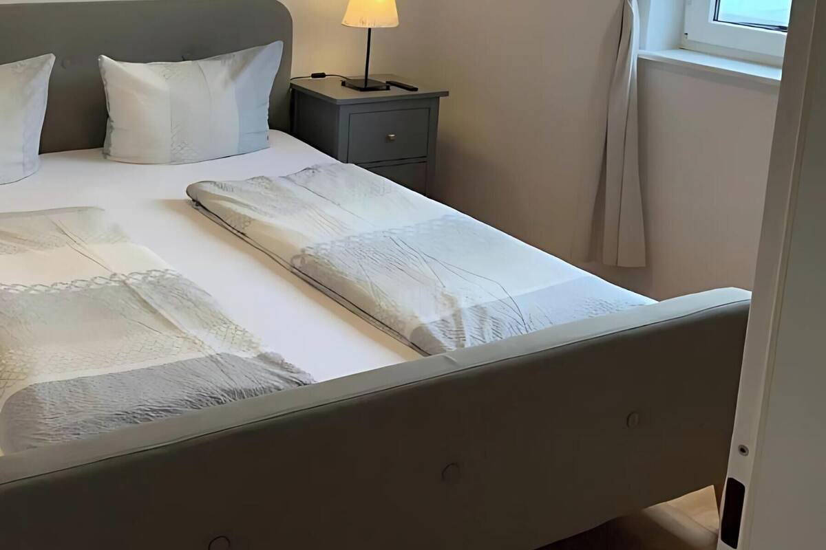 A neatly made bed with a grey headboard, two bedside tables with lamps, and a window with curtains.