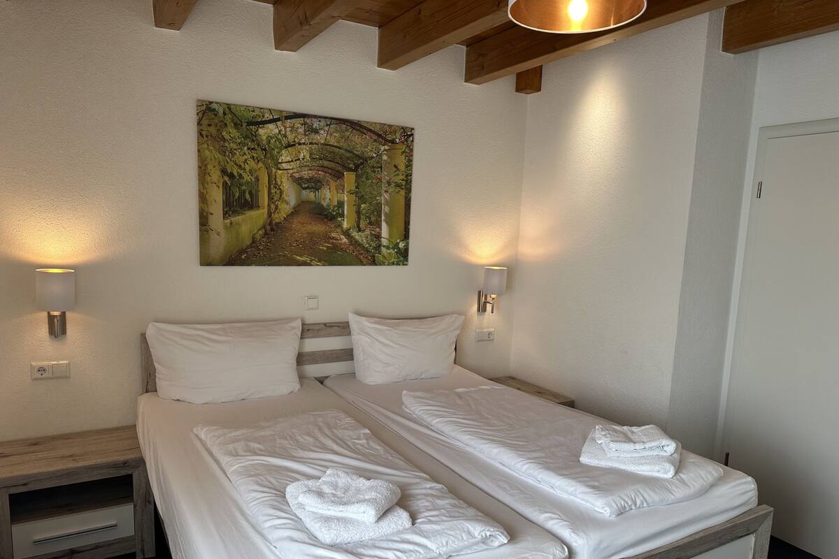 A double bed with white linens and towels, two bedside tables, a painting of a forest path, and wall-mounted lamps.