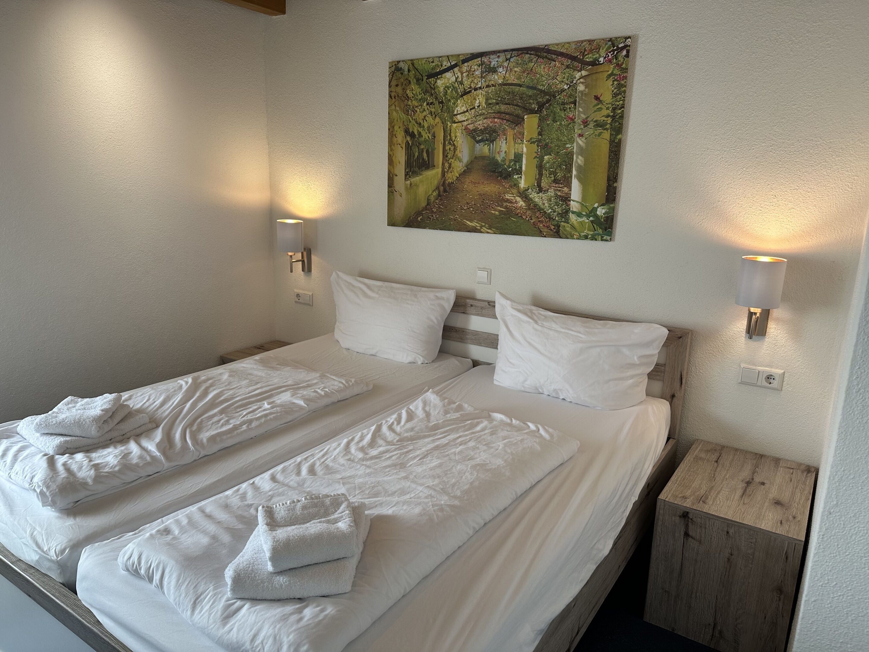 A double bed with white linens and towels, two wall-mounted lights, and a painting of a garden pathway.