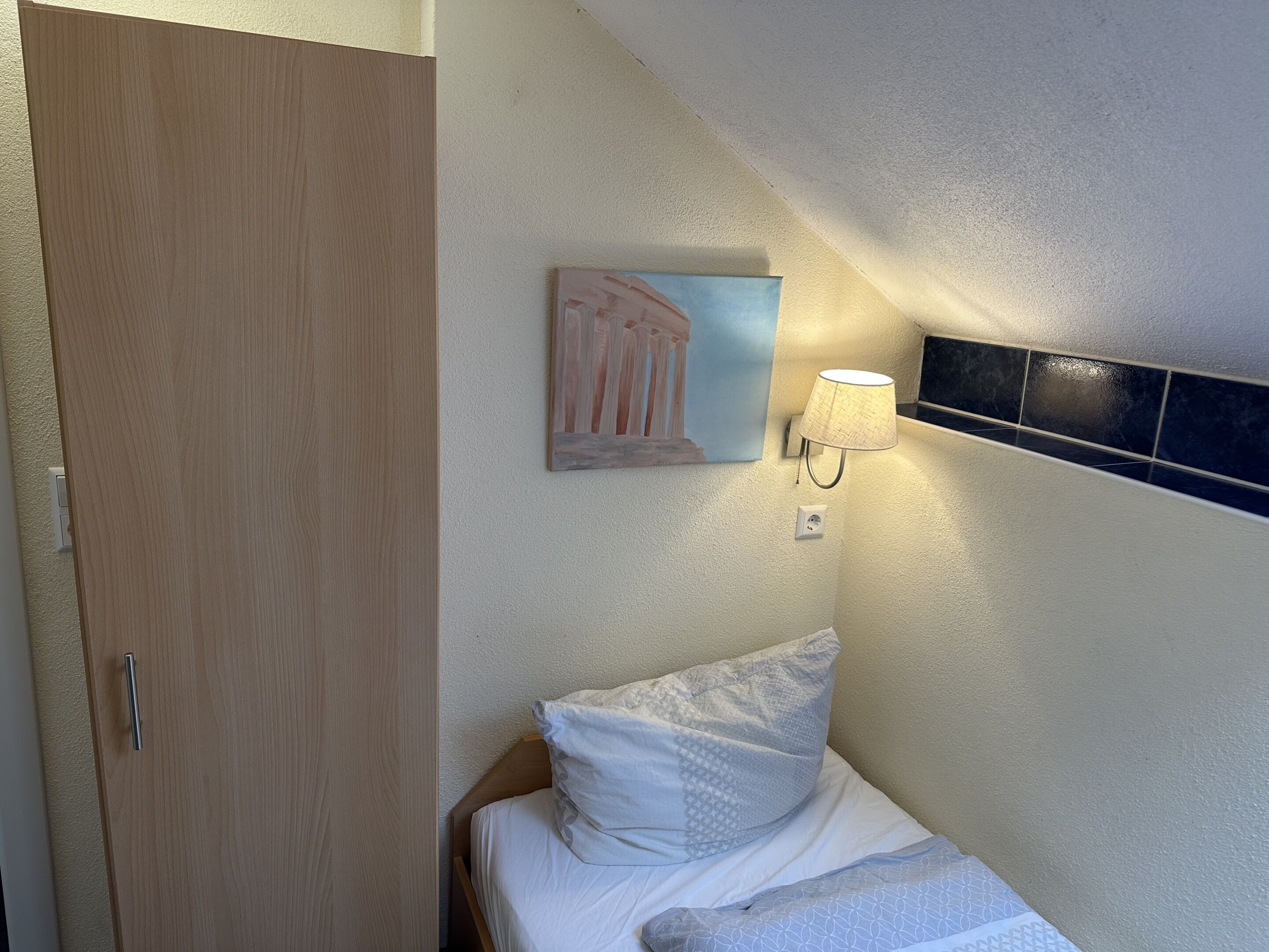 Un dormitorio con puerta de madera, una cama con ropa de cama blanca, una lámpara de pared y un cuadro.