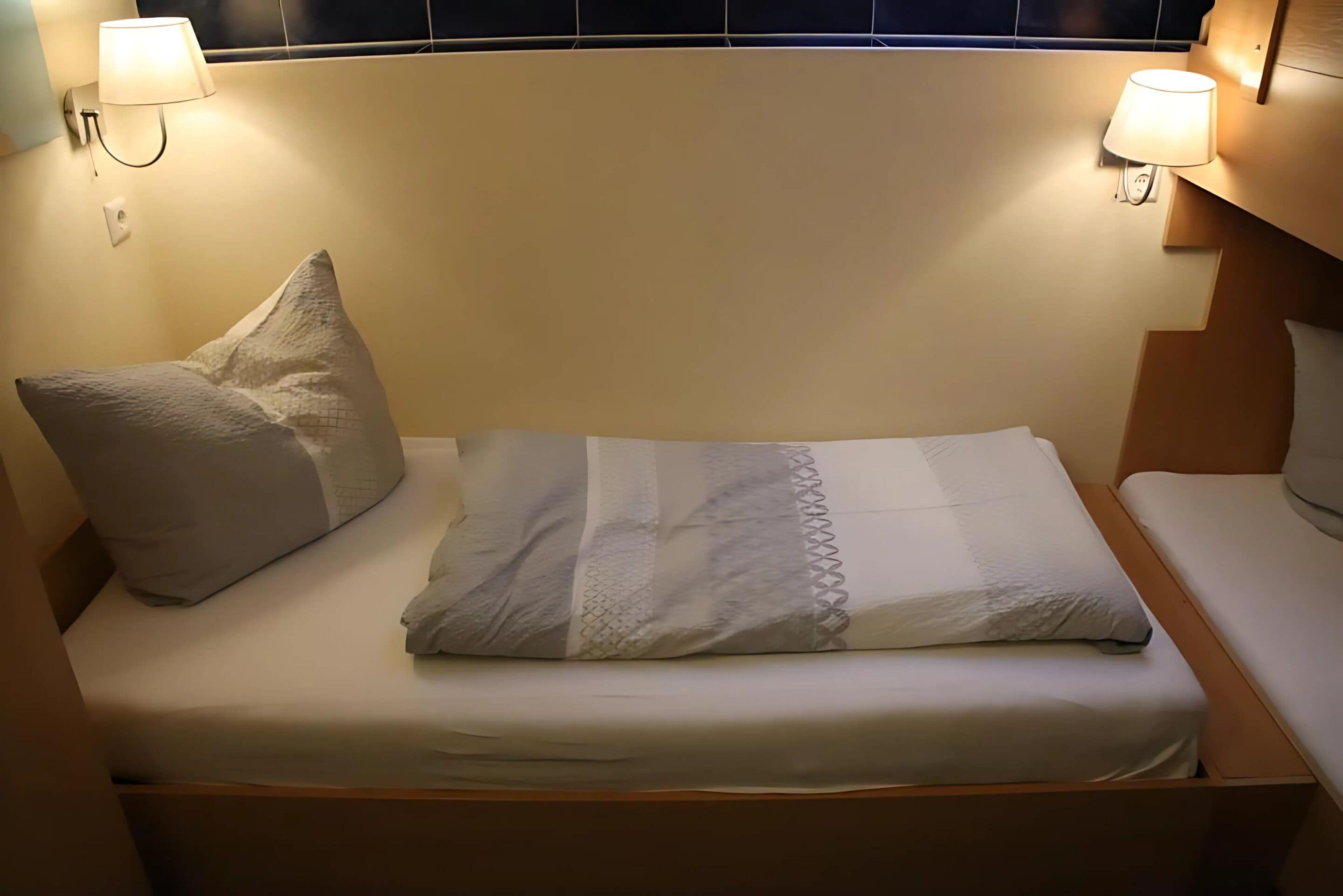 A neatly made bed with a pillow and a blanket, two wall-mounted lamps, and a wooden headboard.
