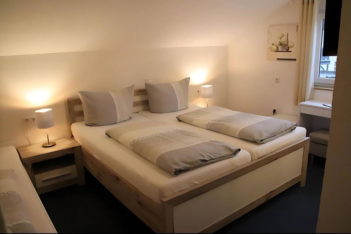 A double bed with two pillows, a bedside table with a lamp, and a small shelf with decorative items.