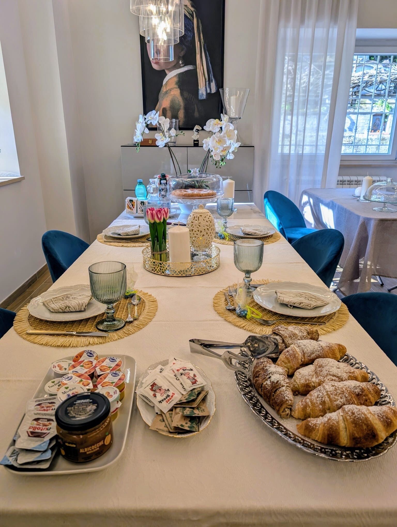 A dining table set with plates, glasses, and pastries, with a decorative centerpiece and a painting in the background.