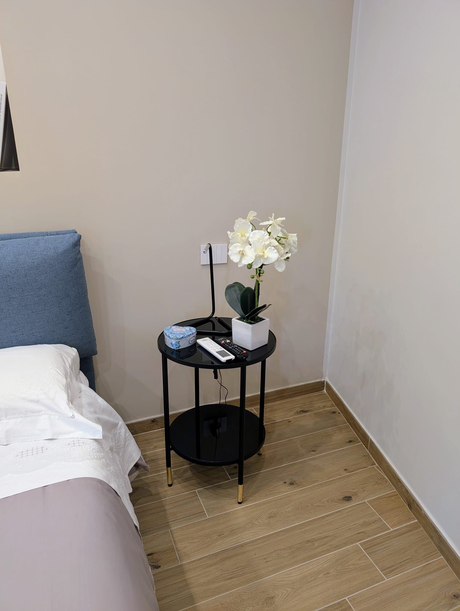 A bedroom with a bed, a small round table with a plant, a remote control, and a decorative box.