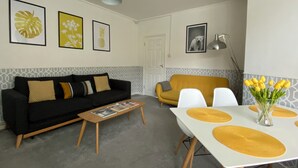 Una sala de estar con un sofá negro, un sofá amarillo, una mesa de comedor con individuales amarillos y una mesa de centro con revistas.