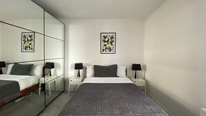 Un dormitorio moderno con un espejo grande, una cama con colcha estampada, dos mesitas de noche con lámparas y dos obras de arte enmarcadas en la pared.