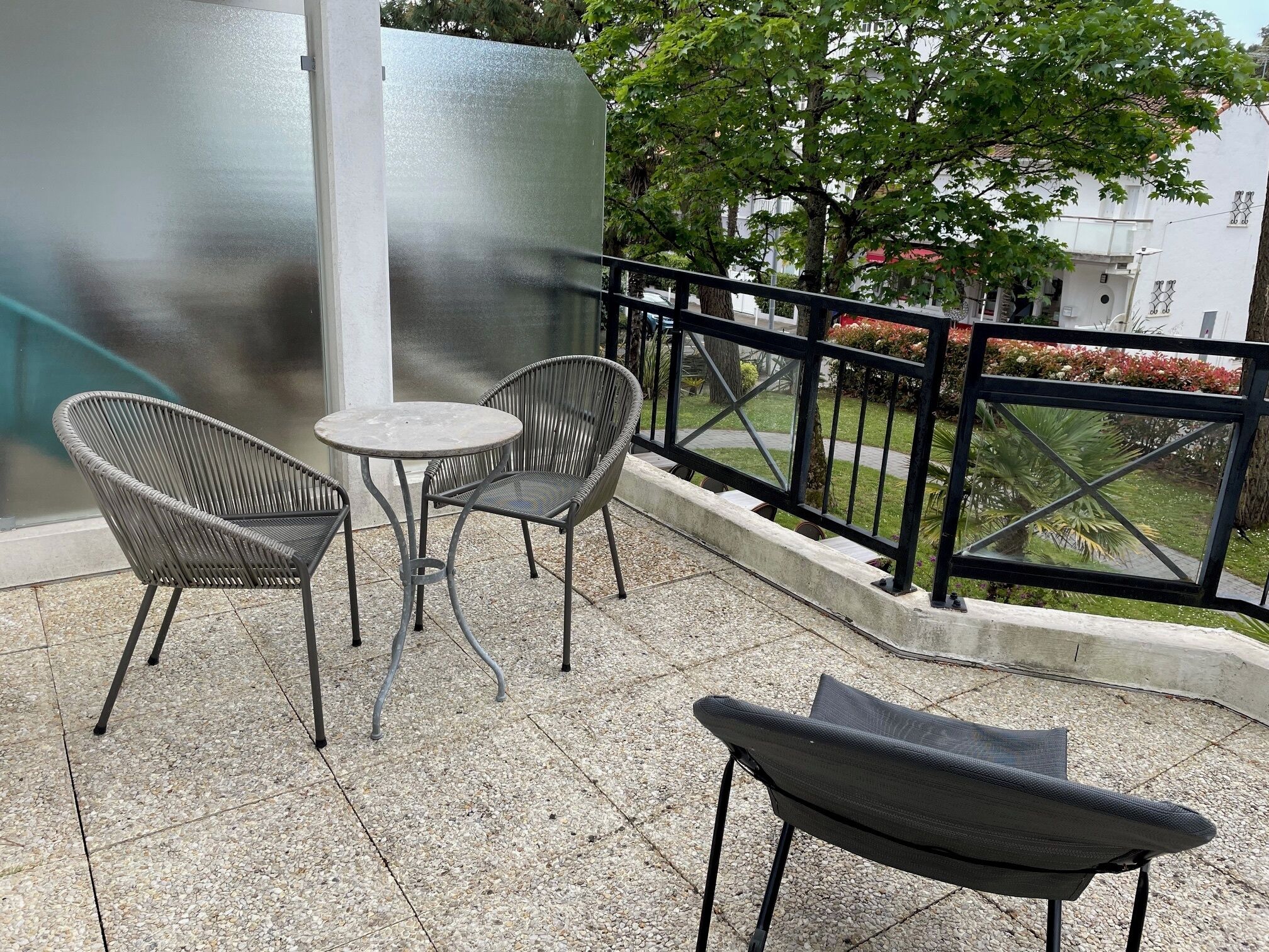 Ein Balkon mit rundem Tisch und zwei Korbstühlen, ein weiterer Stuhl sowie einem Geländer mit Blick auf einen Garten und Gebäude.