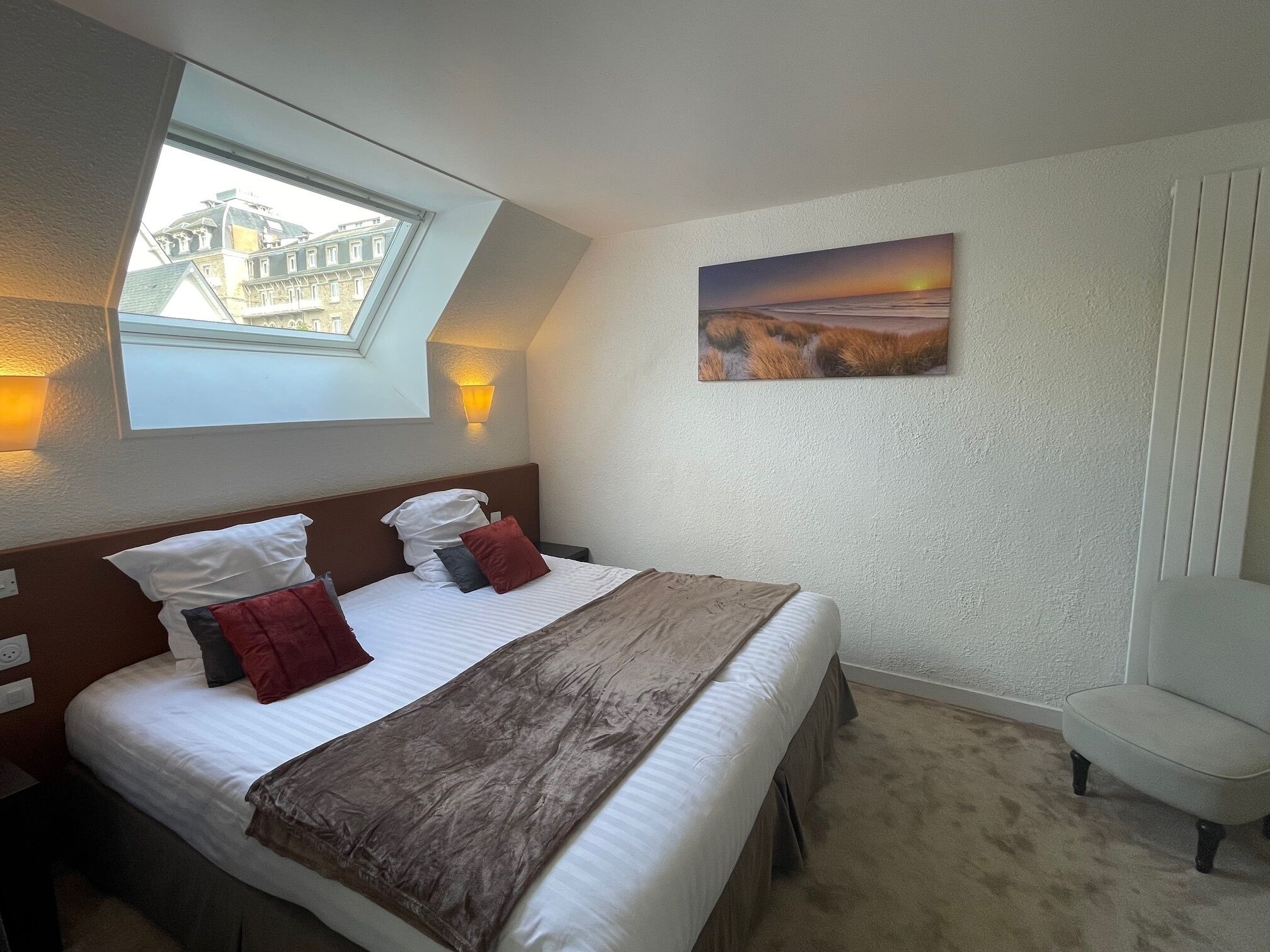A hotel room with a bed, a nightstand, a chair, a painting, and a window with a view of buildings.