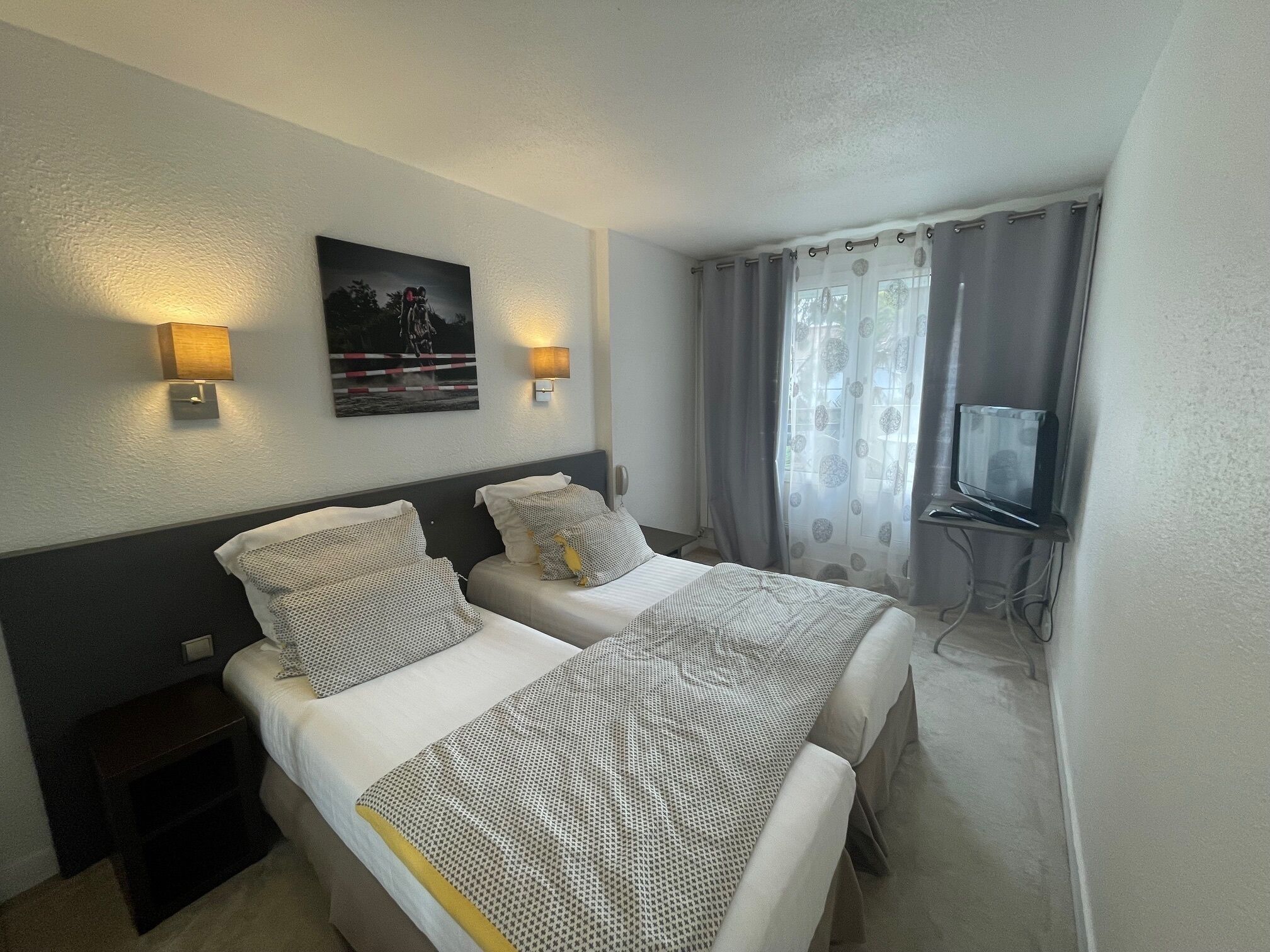 Una camera d'albergo con due letti, un comodino, una lampada a parete, un quadro, un televisore e una finestra con tende.