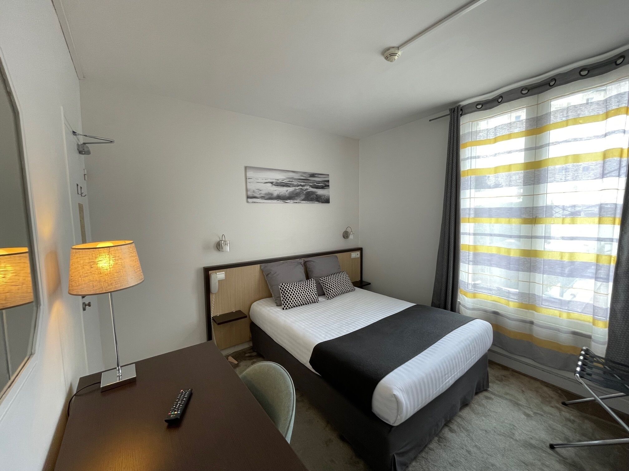 A hotel room with a bed, a desk with a lamp, a chair, a bedside table, and a window with curtains.