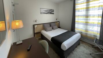 A hotel room with a bed, a desk with a lamp, a chair, a bedside table, and a window with curtains.