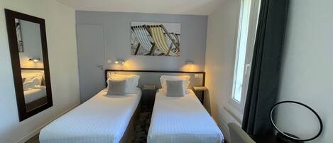 A hotel room with two single beds, a large mirror, a ceiling-mounted smoke detector, a window with curtains, and a wall-mounted artwork.