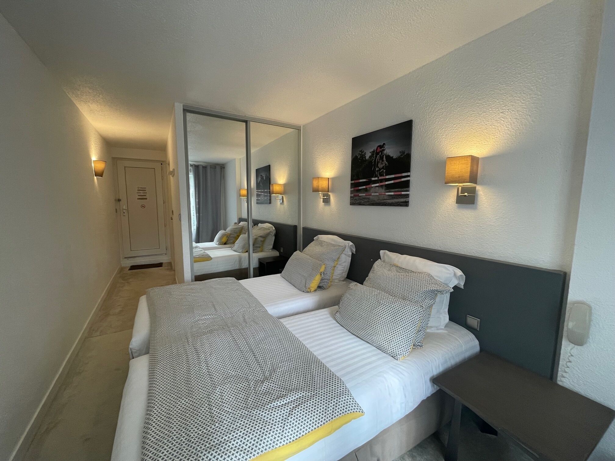 Une chambre d’hôtel avec deux lits, un grand miroir, des lampes fixées au mur et une porte.