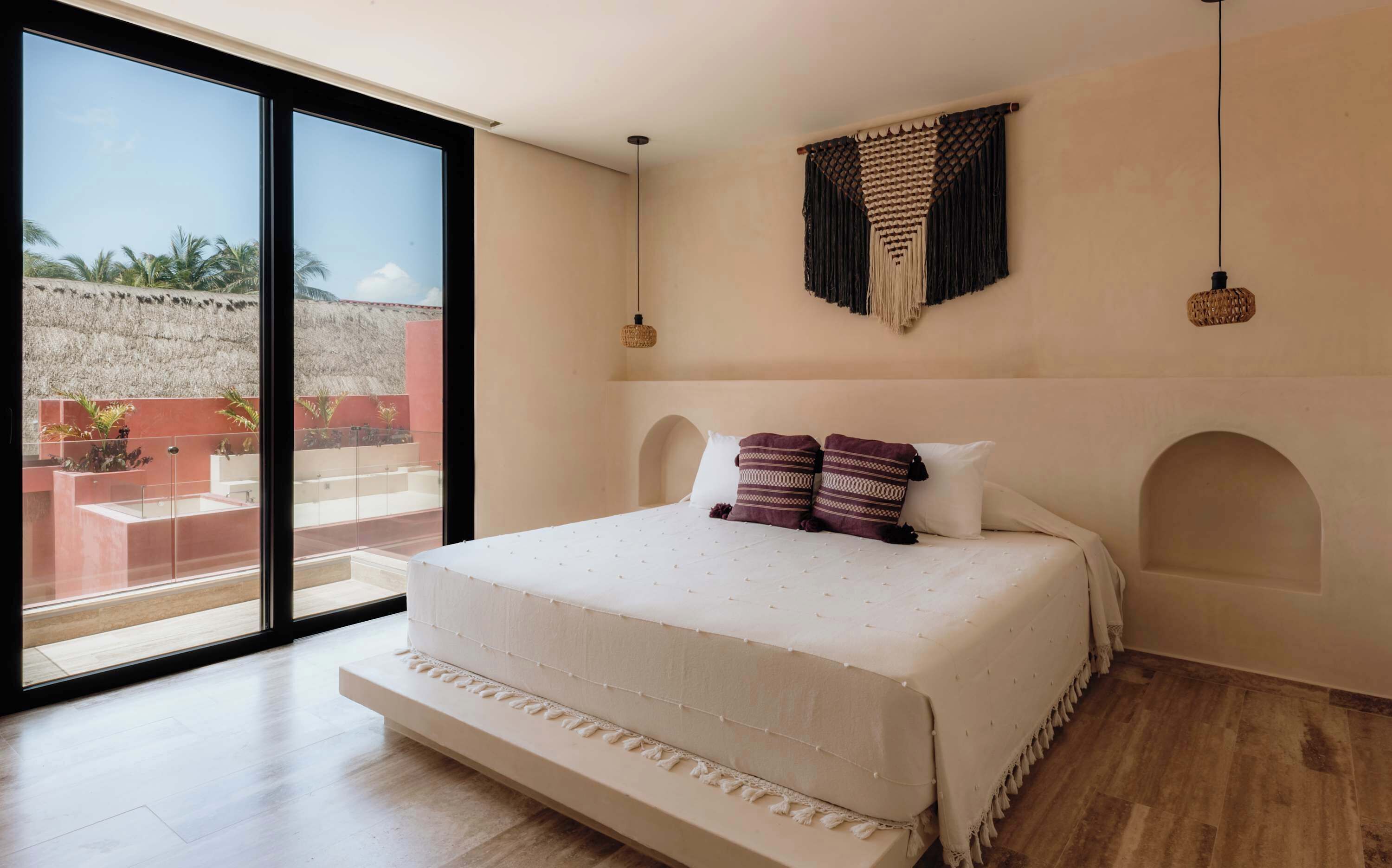 A bedroom with a large window, a bed with a white bedspread, two pillows, and a wall-mounted macrame piece.