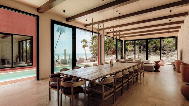 Een ruime eetruimte met een lange houten tafel en meerdere stoelen, grote ramen met uitzicht op het strand en de oceaan, en een modern architectuurontwerp met zichtbare balken en grote glazen deuren.