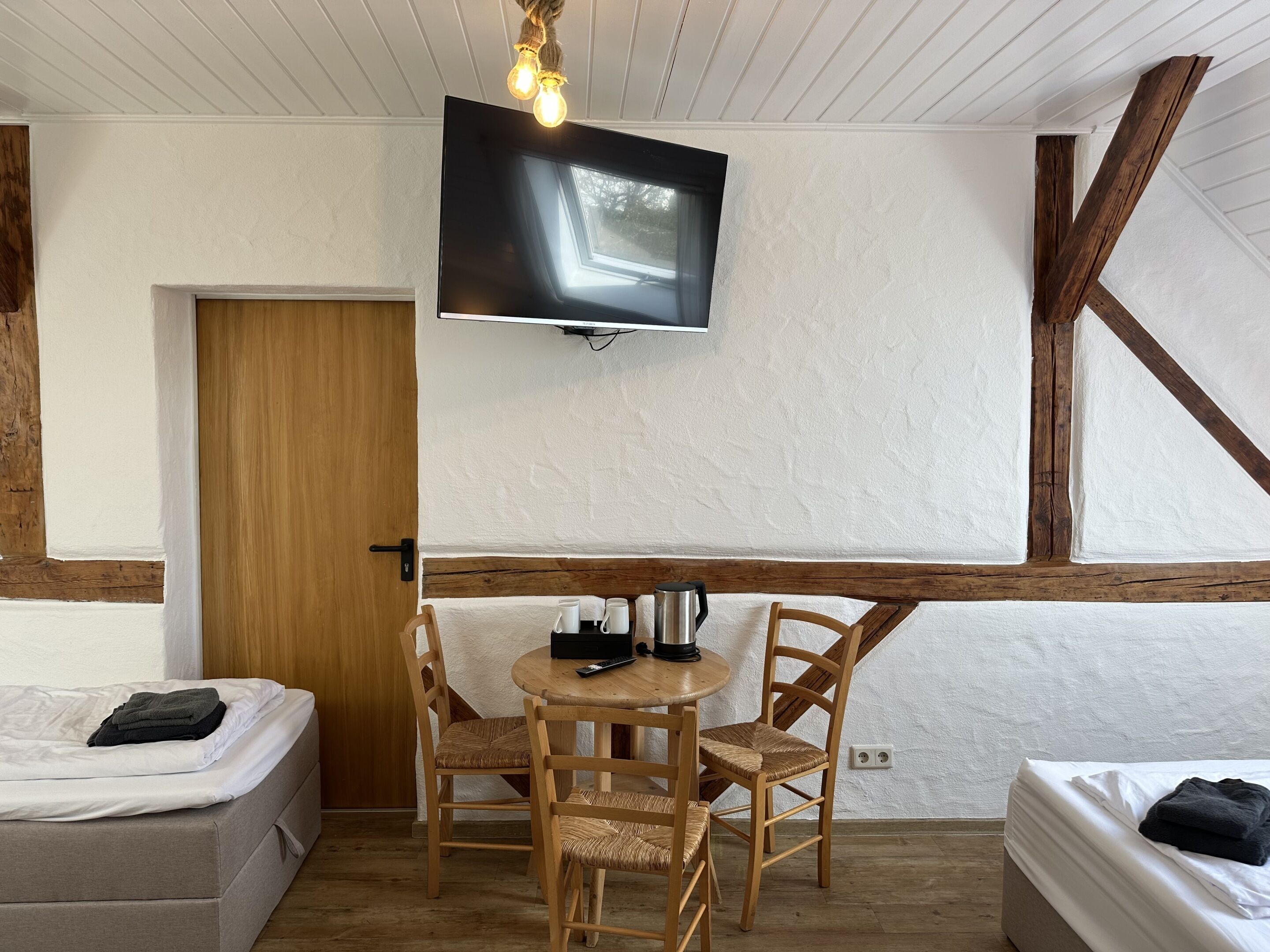 A room with a bed, a table with chairs, a television mounted on the wall, and a wooden ceiling beam.